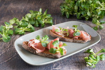 Prosciutto on pieces of bread with greens on hte wooden table.