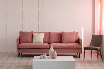 Trendy grey chair next to pastel pink couch with pillows in fashionable living room, copy space on empty wall