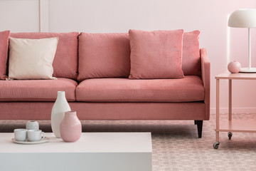 Pastel pink and white vases and coffee mugs on small table in pink living room interior with comfortable sofa