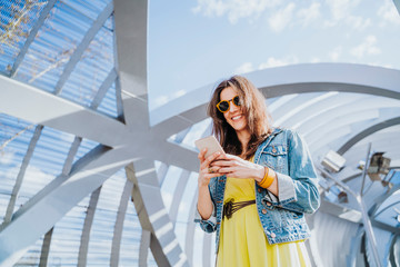 Fototapeta premium Woman wearing yellow shirt texting on the smart phone walking in the street in a sunny day