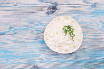 Fresh natural cottage cheese in a white ceramic bowl on the blue wooden table. Green dill. Organic eco healthy meal, dairy product. Top view, overhead photo with place for text.