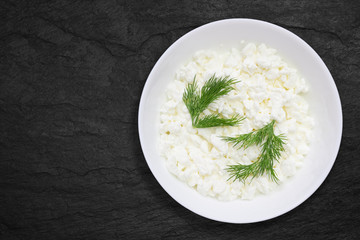 An overhead photo of fresh natural cottage cheese in a white ceramic bowl on the black stone desk. Green dill. Organic eco healthy meal, dairy product. Top view.