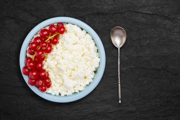 An overhead photo of fresh natural cottage cheese with spoon, red currant in a blue ceramic bowl on the black stone plate. Organic eco healthy meal, dairy product. Top view. Place for text, copyspace.