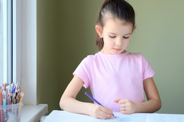 Fototapeta premium Beautiful caucasian white girl with pony tail paints a picture with colorful wooden pencils. Smiling cute girl in pink T-shirt drawing a picture with pencils on blurred background. Child learning art 