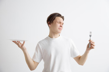 Young sad attractive caucasian guy holding empty dish and fork isolated on grey background. Copy space and mock up. Blank template background. Reject, rejection concept