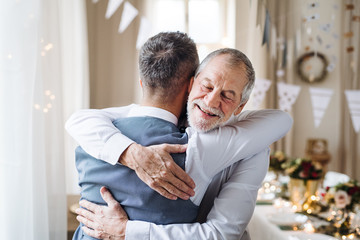 A senior and mature man standing indoors in a room set for a party, hugging.