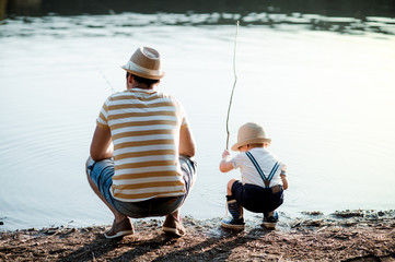 A rear view of mature father with a small toddler son outdoors fishing by a lake.