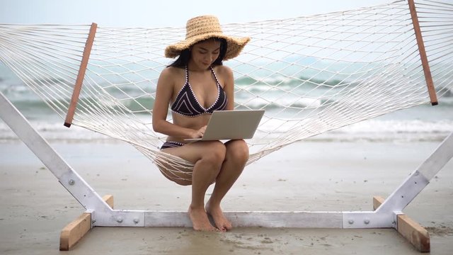 beautiful young asian woman in black bikini and summer hat sitting in hammock working typing with laptop computer on sea beach . girl  sexy working , relaxation , Vacation concept