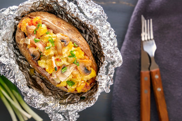 Baked potatoes in foil with bacon, onions and mushrooms with cutlery on a gray wooden table. Close-up. Top view