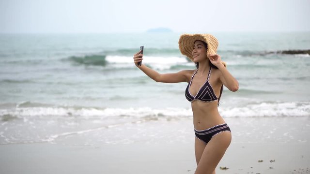 beautiful young asian woman in black bikini and sun hat walking on the sea beach take a picture of herself .summer vacations .girl in  swimsuit  takes selfie photo.