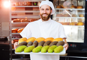 Fototapeta premium young cute Baker in white uniform holding a tray with colored rolls for hot dog on the background of bakery or bread factory