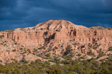 Bright sunlight illuminates a colorful red rock desert peak and badlands under dark, dramatic storm clouds near Santa Fe, New Mexico