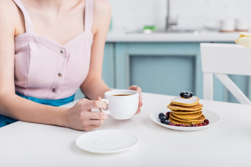 cropped view of young woman holding cup of coffee near plate with pancakes on table