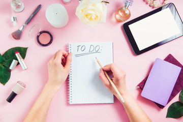 Female hands write To do list on the pink background with cosmetics, coffee cup, notebooks, tablet with blank screen. Beauty blog concept. Top view feminine office table desk. Freelancer working place