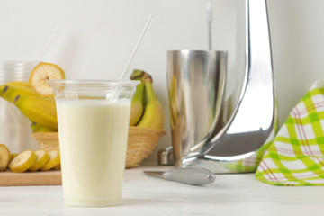 Making a milkshake. plastic disposable glass with a banana milkshake, ingredients for cooking and mixer on a light background