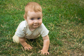 Little toddler boy trains his crawling skills. The child happily crawls on the green grass. Toddler smiles and moves on all fours around the yard in the open air.