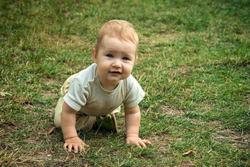 Little toddler boy trains his crawling skills. The child happily crawls on the green grass. Toddler smiles and moves on all fours around the yard in the open air.