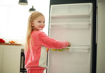 Joyful girl child near refrigerator holding an apple in hands, healthy food as a lifestyle