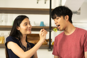 Asian young couple is feeding each other and smiling while cooking salad in kitchen