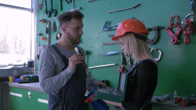 internship in car service station, professional mechanic teaches young girl into helmet with working set of tools for repair automobile at workshop