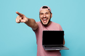 Trendy hipster guy with beard wearing pink outfit and cap holding new laptop in hands and pointing away on blue background