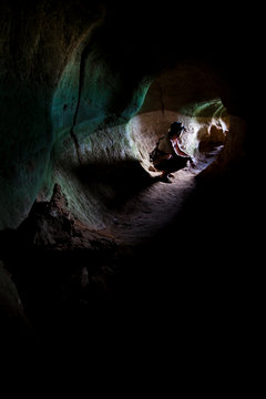 Italian male caver squating in a limestone tunnel in Krabi Province, Thailand.  The subject is looking out to the exit/entrance.  The light source is at the mouth of the cave and backlights the subject heavily.  This image includes a lot of black space because of the extreme lighting situaton.  Shot in the early afternoon in the late summer.