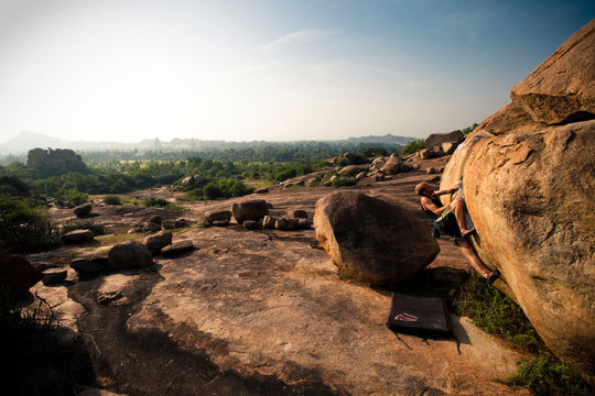 A solo, caucasian, male climber pulls onto his project boulder in Hampi, India.  Other than scattered bushes, the landscape is bare stone.  The horizon fades off into early morning haze in the distance.  Shot in the morning in Autumn (Fall).
