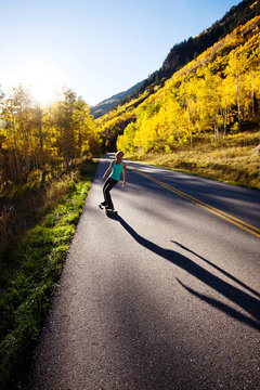 A young woman longboards down a smooth country road through the mountain peaks of Aspen at the peak of fall colors.