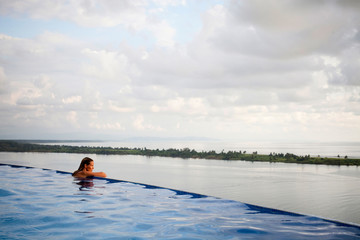 A woman sits with her arms on the edge of an infinity pool overlooking the wide open ocean.