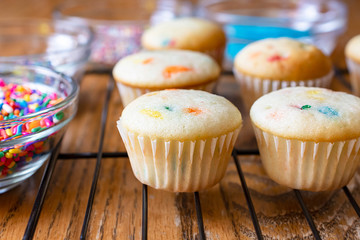 Mini cupcakes on wire rack waiting to be frosted.  Various colored sugars and sprinkles in glass bowls around cupcakes.