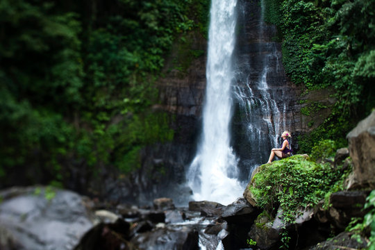 A beautiful young woman sitting in a lush jungle next to a waterfall in Lovina, Bali, Indonesia.