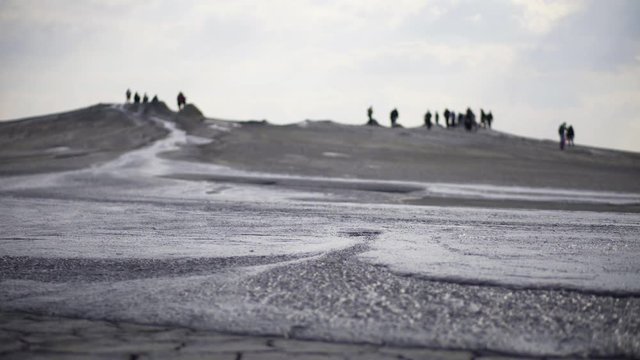 Stream of volcano mud flowing on a dried cracked soil. Tourists walking next to mud volcanoes craters on a windy cloudy day. Buzau county, Romania