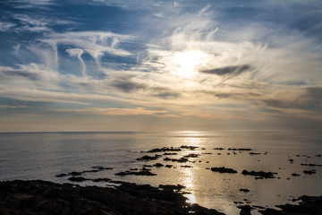 Blue sky at dawn covered by strange cloud formations on a flat sea with black rocks that stand out in the reflection of the sun on the water