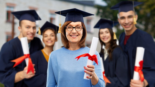 Graduation, Education And Old Age Concept - Happy Senior Graduate Student Woman In Mortar Board With Diploma Next To Young People Over University Campus Background