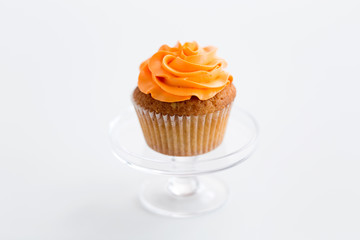 food, pastry and sweets concept - cupcake with orange buttercream frosting on glass confectionery stand over white background