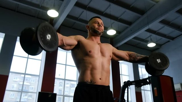 Bodybuilder makes distributing of hands with dumbbells while standing in the gym, maintaining a healthy lifestyle