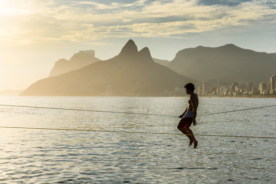 A Man Sitting On A Slackline At Arpoador Beach During The Sunset, Rio De Janeiro, Brazil
