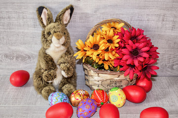 Colorful painted Easter eggs lie in a basket with flowers, on a wooden background, with a fluffy Easter bunny.