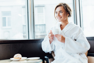 attractive brunette woman in bathrobe smiling and holding cup of coffee