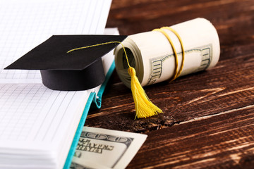Small graduation cap with dollar banknotes and notepad on brown wooden table
