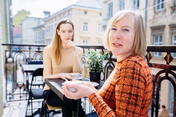 Family day. Middle age blond woman in orange dress and her adult daughter drinking coffee at balcony of cafe or home with city view on background. Relationships and shared news family time concept.