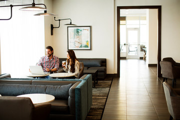 A young couple sits side by side on two sofas, looks at a laptop that is on one of two round shape table before them and enjoys some soft drink.