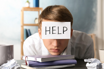 Young man with note HELP on forehead at workplace