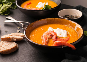 pumpkin soup with shrimps, sour, pumpkin seeds in dark bowls and bread, greenery, silver spoons on black background