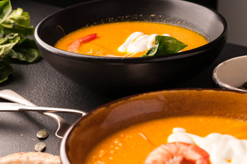 pumpkin soup with shrimps, pumpkin seeds in dark bowls and bread, silver spoons on black background