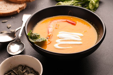 pumpkin orange soup with shrimp, sour in dark bowlon and bread near silver spoons on black background