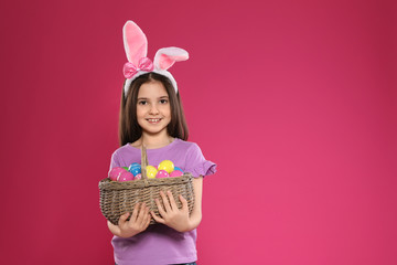 Little girl in bunny ears headband holding basket with Easter eggs on color background, space for text