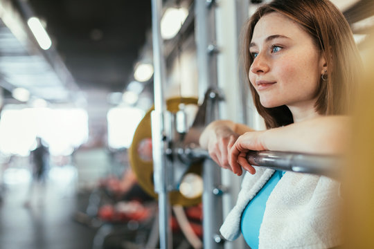 Young woman exercising in gym room. Nice cheerful model lean to barbell in smith machine and smile. Resting. Towel around neck. Alone in fitness room.