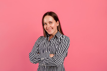 Nice confident young woman in striped shirt pose on camera and smile. Hands crossed. Positive mood and happy. Isolated over pink background.