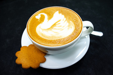  Cappuccino with swan drawn on the foam, in a white cup with spoon, saucer and biscuit, black background